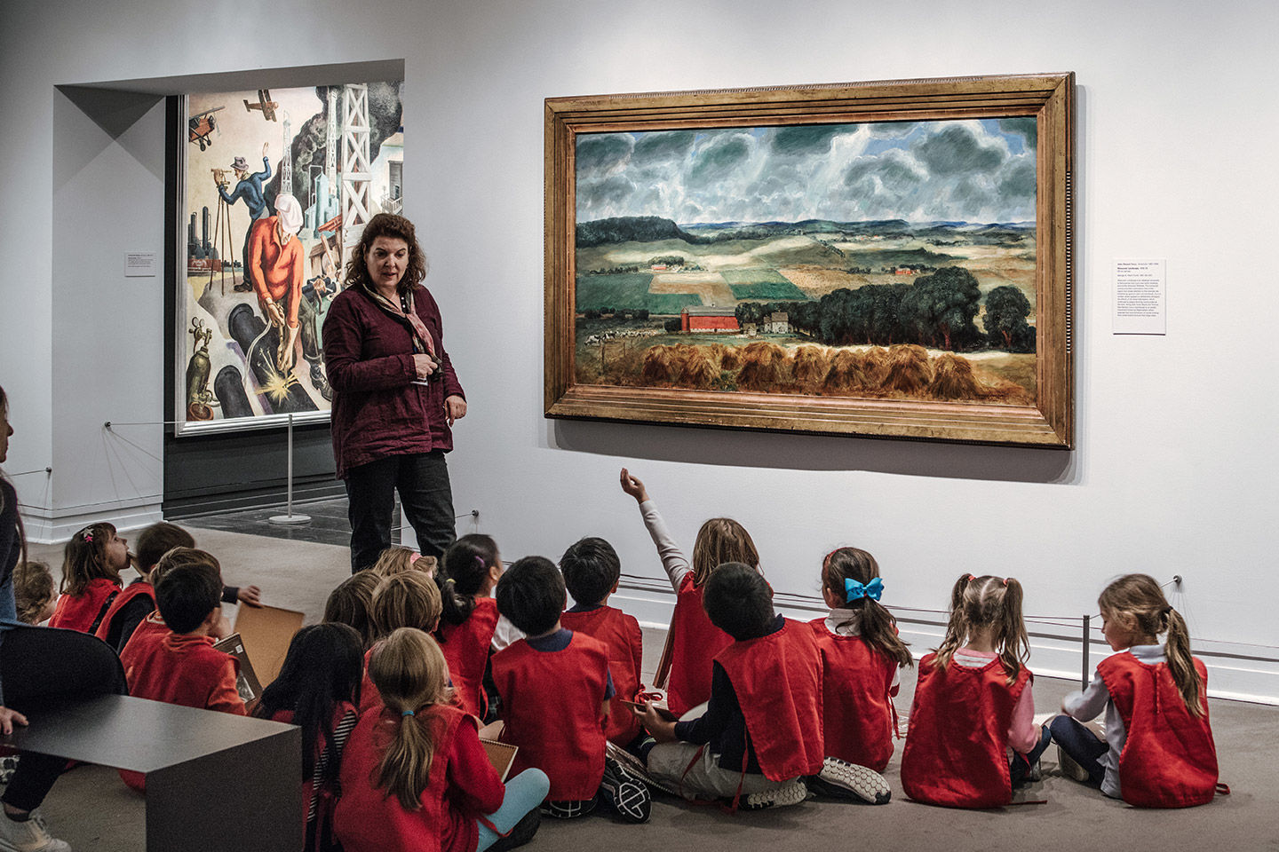 A group of children wearing uniform red vests sit on the floor of an art gallery and look up to a woman talking to the side of a large landscape painting of farmland, hills, and sky filled densely with animated clouds